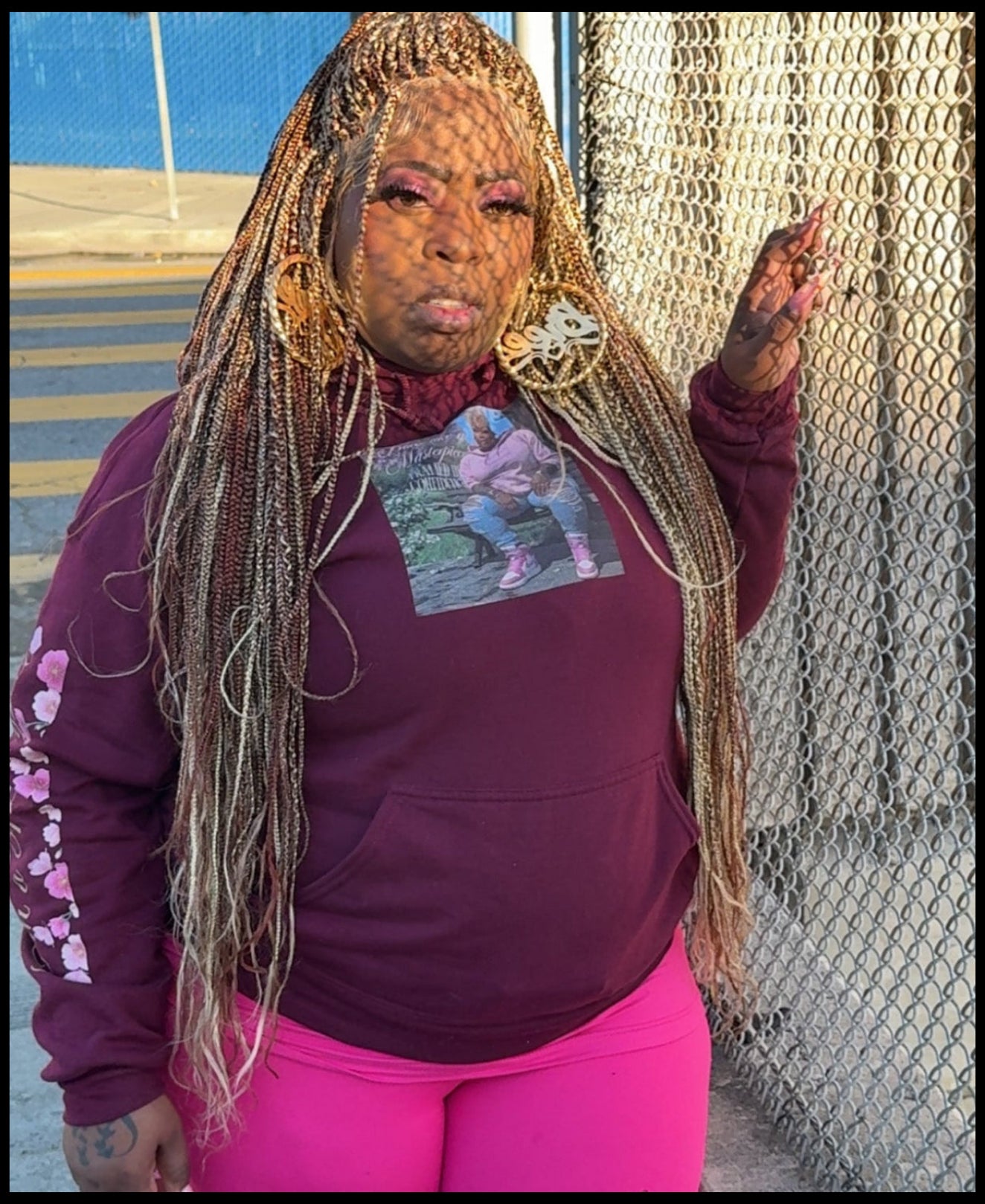 Person wearing a maroon hoodie and pink pants standing next to a chain-link fence.